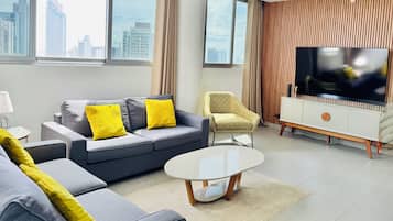 Una sala de estar moderna con un sofá gris, cojines amarillos, una mesa de centro redonda, un sillón estampado y un televisor grande sobre un mueble de madera.