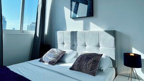 A modern hotel room with a large bed, a nightstand with a lamp, a window with curtains, and a city view.