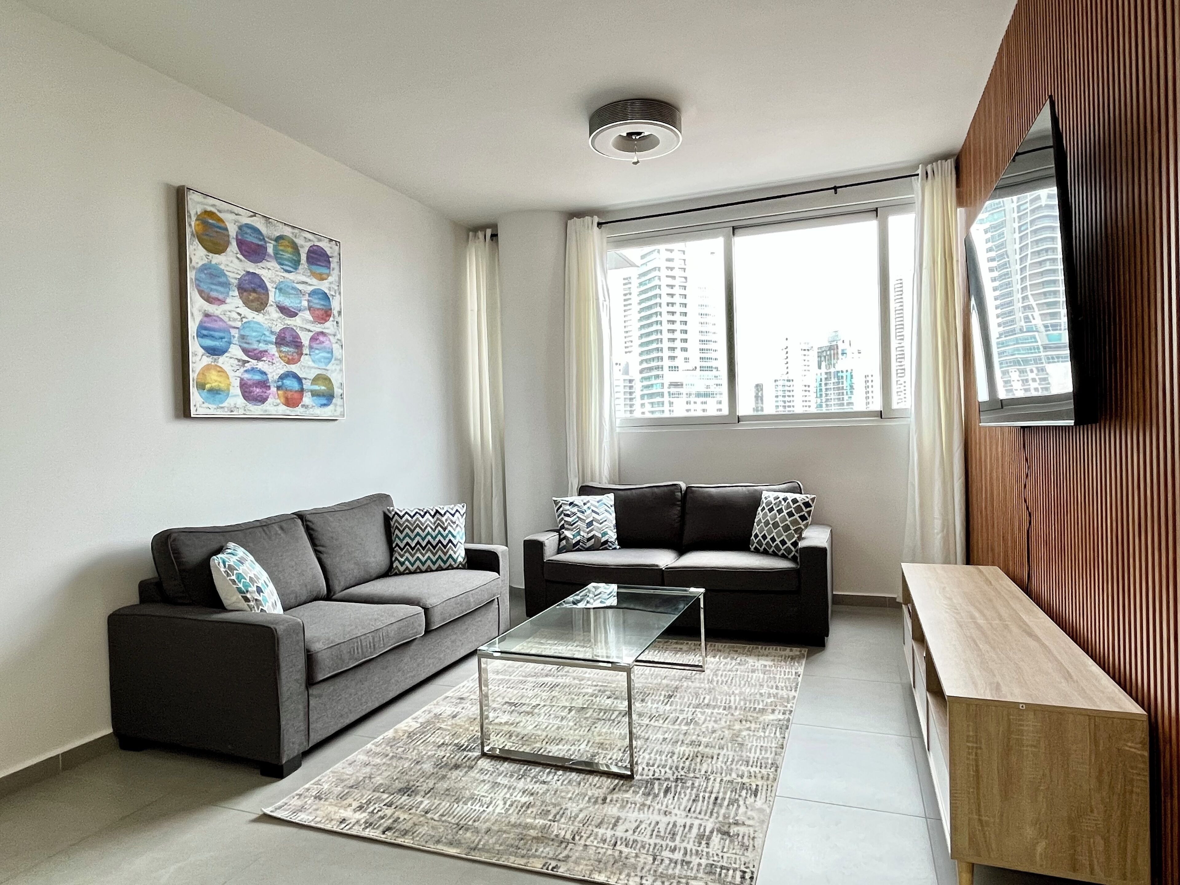 Een moderne woonkamer met twee banken, een glazen salontafel, een houten bijzettafeltje, uitzicht op de stad door grote ramen en een plafondlamp.