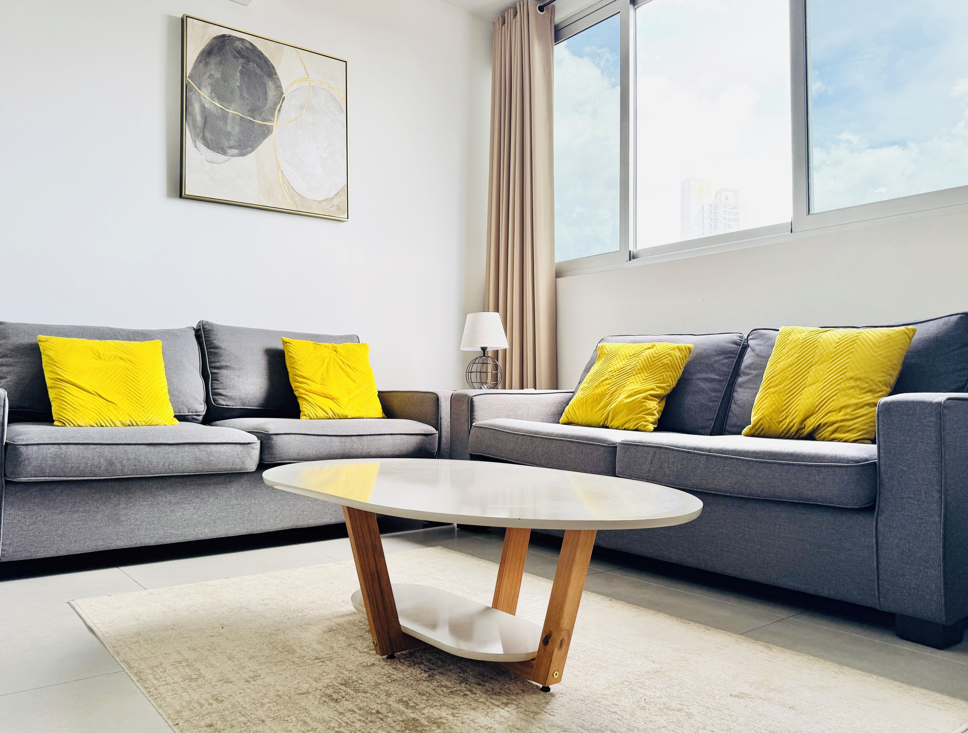 A modern living room with two grey sofas, each with a yellow cushion, a round coffee table with wooden legs, and a floor lamp.