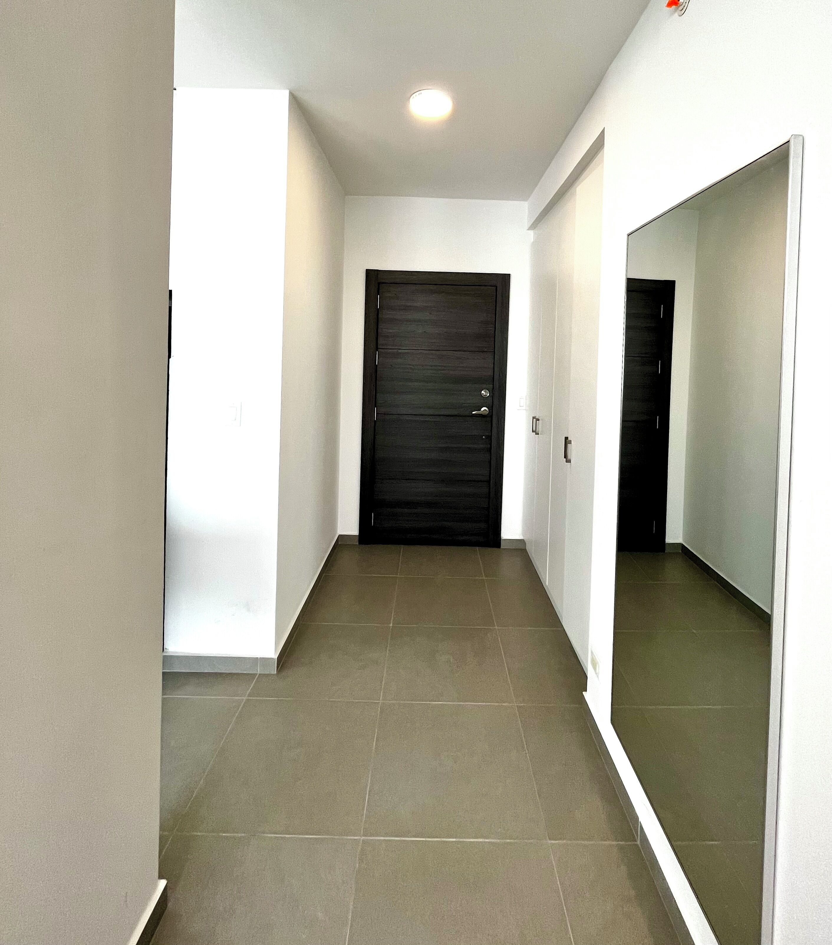 A hallway with a dark door, white walls, and tiled flooring.