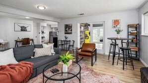 A modern living room with a grey sofa, a brown leather chair, and a coffee table with a plant. There is a dining area with a small table and bar stools. - Arlington Retreat Near Stadiums and Six Flags (Arlington)