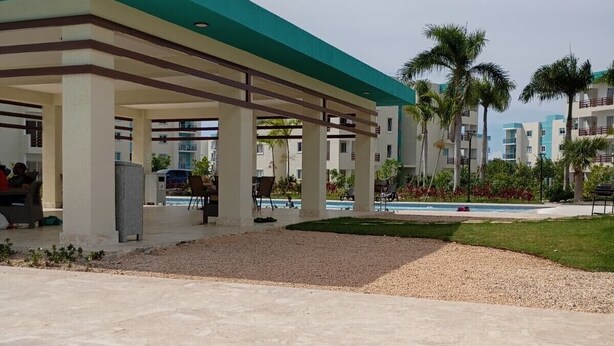 A covered outdoor area with a pool, palm trees, and a building with a green roof. - Charming  pool view apartment at Serena village, with Beach access in Punta Cana (Punta Cana)