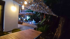 Un área de comedor al aire libre con mesas y sillas de madera, rodeada de plantas tropicales y una sombrilla de techo de paja.