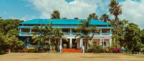 A two-story building with a blue roof and white columns, surrounded by palm trees and a grassy lawn.