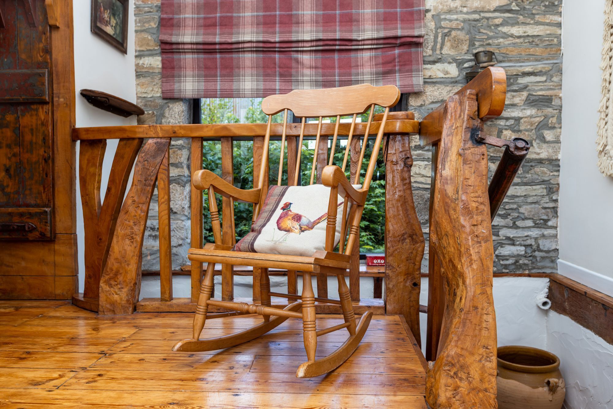 A wooden rocking chair with a cushion, positioned in a room with a stone wall and a window with a plaid curtain.