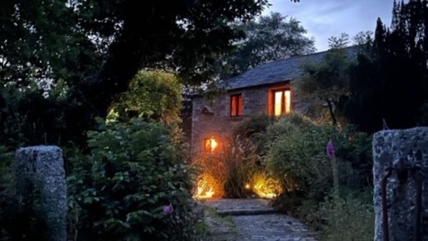 Un camino de piedra que conduce a una casa rústica con luz en su interior, rodeada de vegetación.