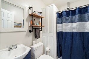 A bathroom with a white sink, a toilet, a mirror, a shelf with toiletries, and a blue striped shower curtain. - Charming 2-bedroom barn in, Foley 8 miles to Beach, dogs welcome, near Sportplex (Foley)