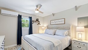 A bedroom with a bed, bedside tables, a ceiling fan, an air conditioning unit, a window with curtains, and a wall with a framed sign. - Charming 2-bedroom barn in, Foley 8 miles to Beach, dogs welcome, near Sportplex (Foley)