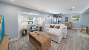 A spacious living room with a white sofa, a blue armchair, and a wooden bench. There are two windows with blinds, a lamp on a side table, and a spiral staircase in the background. - Fresh Coastal Comfort, Beaches Close & Bay Views, 2 GB WIFI (Sarasota)