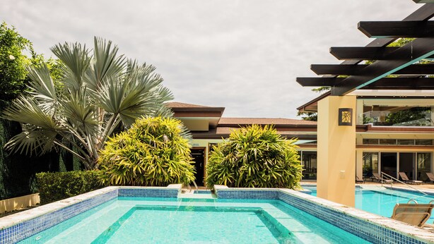 A swimming pool with tiled edges, surrounded by lush greenery and a modern building with a sloped roof.