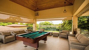 A pool table in a sunlit room with wicker furniture and large windows.
