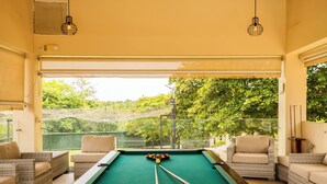 A pool table in a room with wicker furniture and a view of greenery outside.