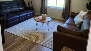 Una sala de estar con un sofá de cuero negro, un sofá de cuero marrón, una mesa de centro redonda, una silla de mimbre, una lámpara de pie y una ventana con cortinas.