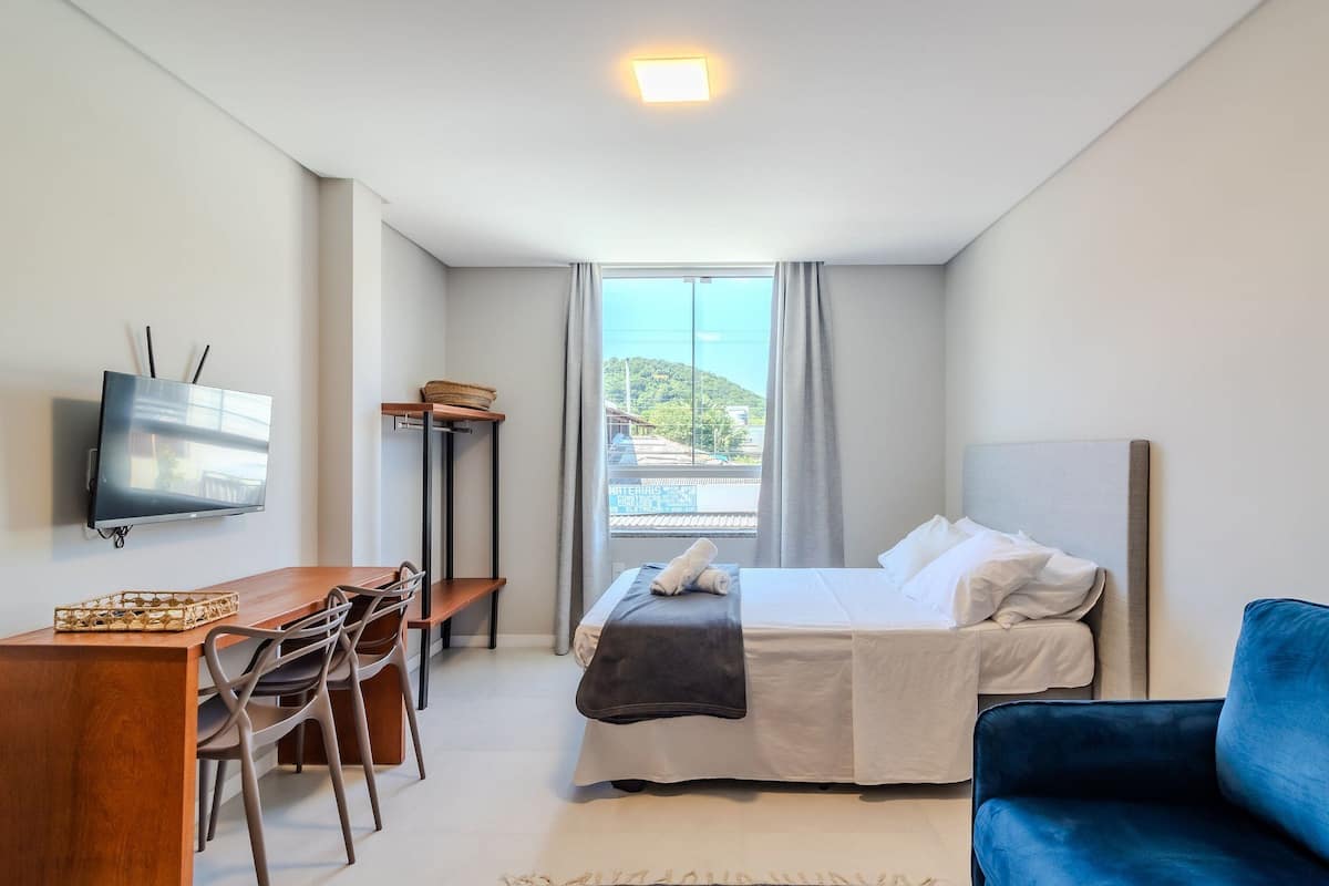 Ein Hotelzimmer mit einem Bett, einem Schreibtisch mit Stühlen, einem Fernseher und Blick auf ein Gebäude und Hügel.