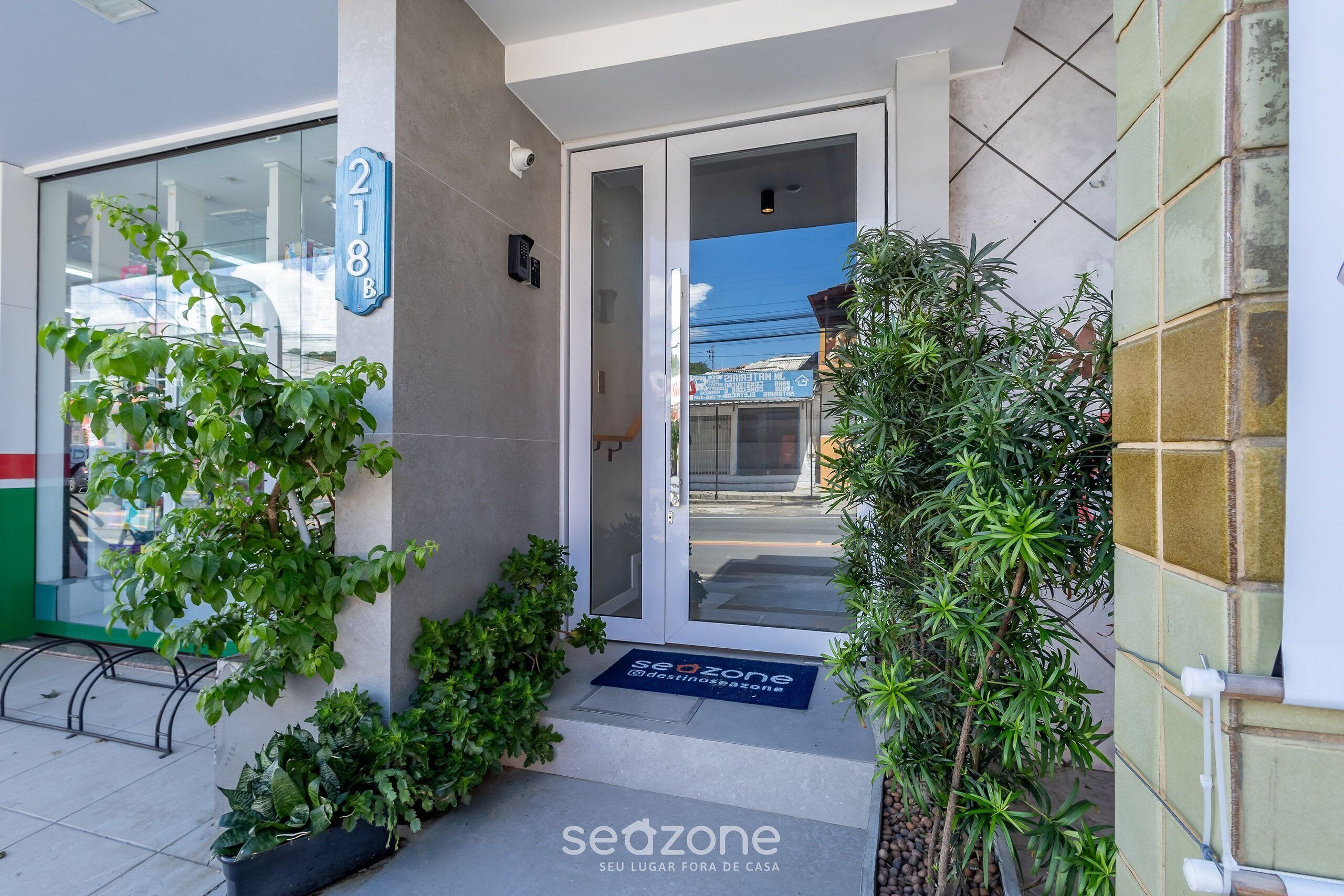 A building entrance with a glass door, a number sign '218B', and a doormat with 'sedzone' branding.