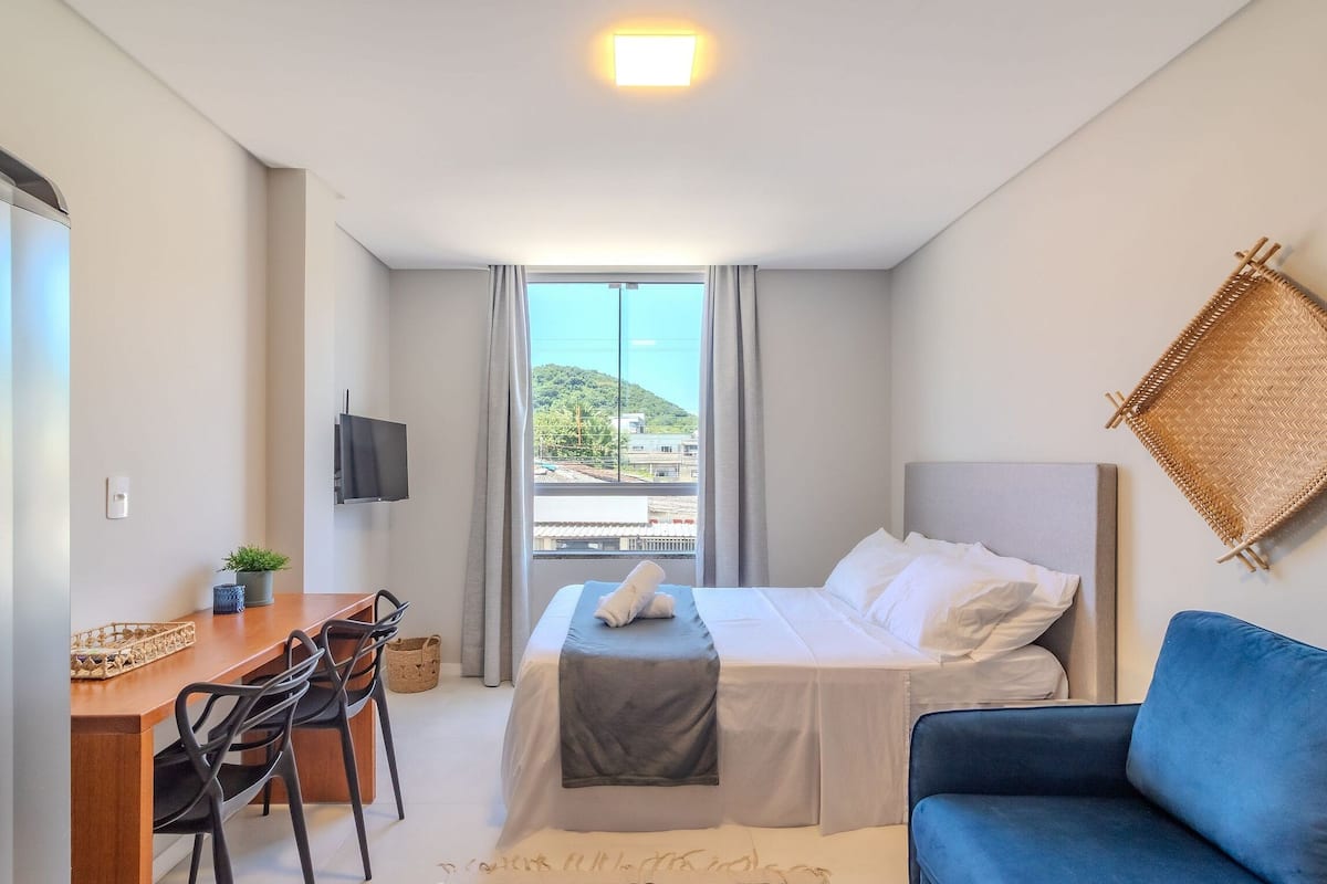 Ein Hotelzimmer mit Bett, Schreibtisch, Stuhl, blauem Sofa, Fernseher und Fenster mit Blick auf Gebäude und Hügel.