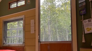 Una cabaña con una cama litera, una ventana que da a los árboles y una puerta que conduce al exterior.