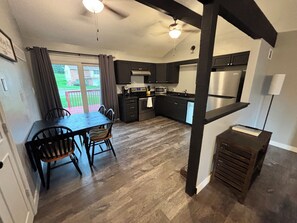 Une cuisine moderne avec des meubles de couleur sombre, un coin repas avec une table et des chaises en bois, et une vue sur un espace extérieur verdoyant grâce à de grandes fenêtres.