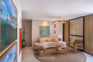 A modern living room with a curved sofa, a wooden coffee table, and a chair. There is a painting on the wall and a potted plant on the floor. - BLB - Apartments in Barra, Salvador (Salvador)