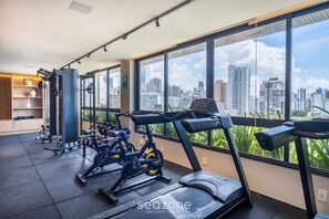 A gym with exercise equipment, large windows, and a city view. - BLB - Apartments in Barra, Salvador (Salvador)