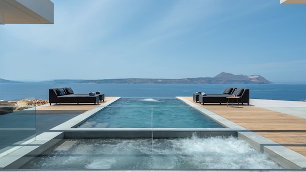 A modern infinity pool with a wooden deck, two black lounge chairs, and a clear view of the sea and mountains in the background.