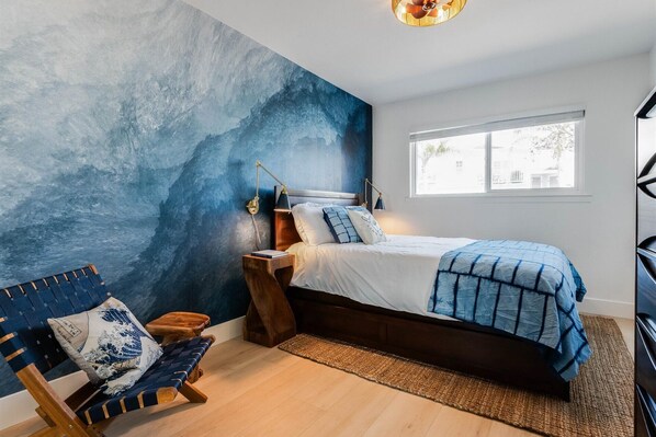Ein Schlafzimmer mit einer großen, blauen abstrakten Wandmalerei, einem Bettgestell aus Holz, einem Nachttisch mit Lampe, einem Sessel mit blauem Kissen und einem Fenster mit Blick auf Gebäude.