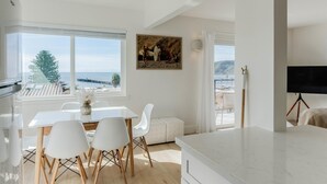 Una cocina moderna con un ventanal que da a la costa, un comedor con sillas blancas y un cuadro en la pared.