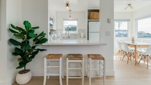 Una cocina moderna con armarios blancos, un espejo grande y vista al área de comedor. Hay tres taburetes blancos con asientos de mimbre y una planta grande en maceta.