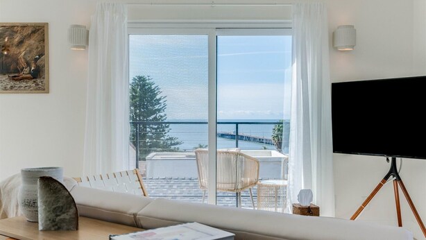 Una habitación moderna con un ventanal grande con vista a una masa de agua, un televisor de pantalla plana, un sofá y una pequeña mesa con libros.