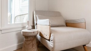 Habitación de esquina con un sillón de color claro, una pequeña mesa redonda con un objeto blanco y un espejo circular en la pared.