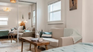Una sala moderna con un sofá, una mesita de centro y una cama. Hay un espejo grande, una ventana y un cuadro en la pared.