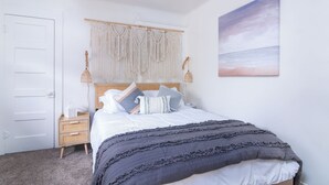 Un dormitorio con una cama, un tapiz de macramé, una mesita de noche, una silla de mimbre y un cuadro en la pared.