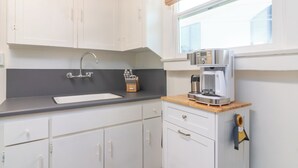 Una cocina con armarios blancos, encimera negra, cafetera de acero inoxidable y una ventana con persiana de bambú.