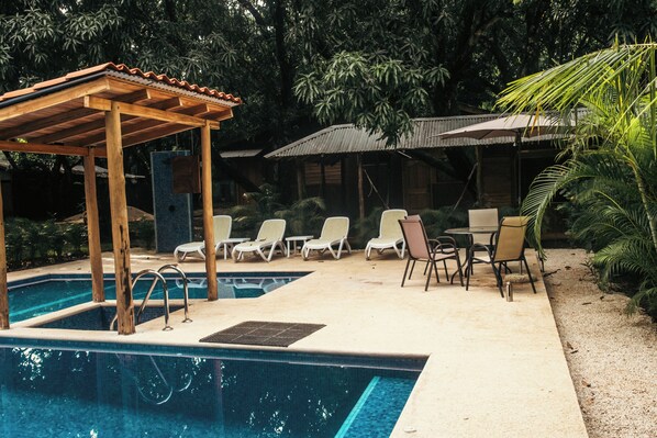 A pool area with two pools, lounge chairs, a pergola, and a covered seating area.