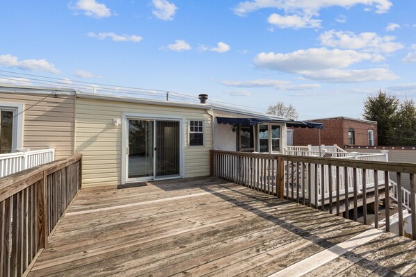 Une terrasse en bois avec une porte vitrée donnant sur un espace abrité, des rampes blanches et un bâtiment en briques en arrière-plan.