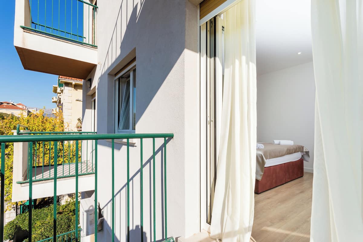 Un balcone con ringhiera verde, un letto con copriletto bianco e marrone, e una finestra con tende bianche.