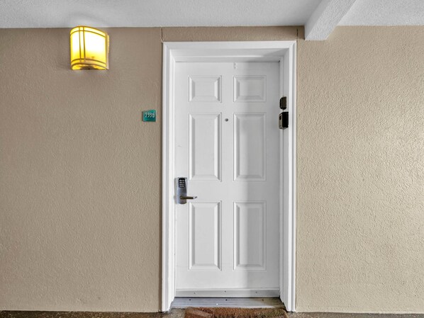 Une porte blanche dotée d’un clavier pour l’ouverture, une plaque verte indiquant le numéro de la maison et un luminaire jaune fixé au mur.