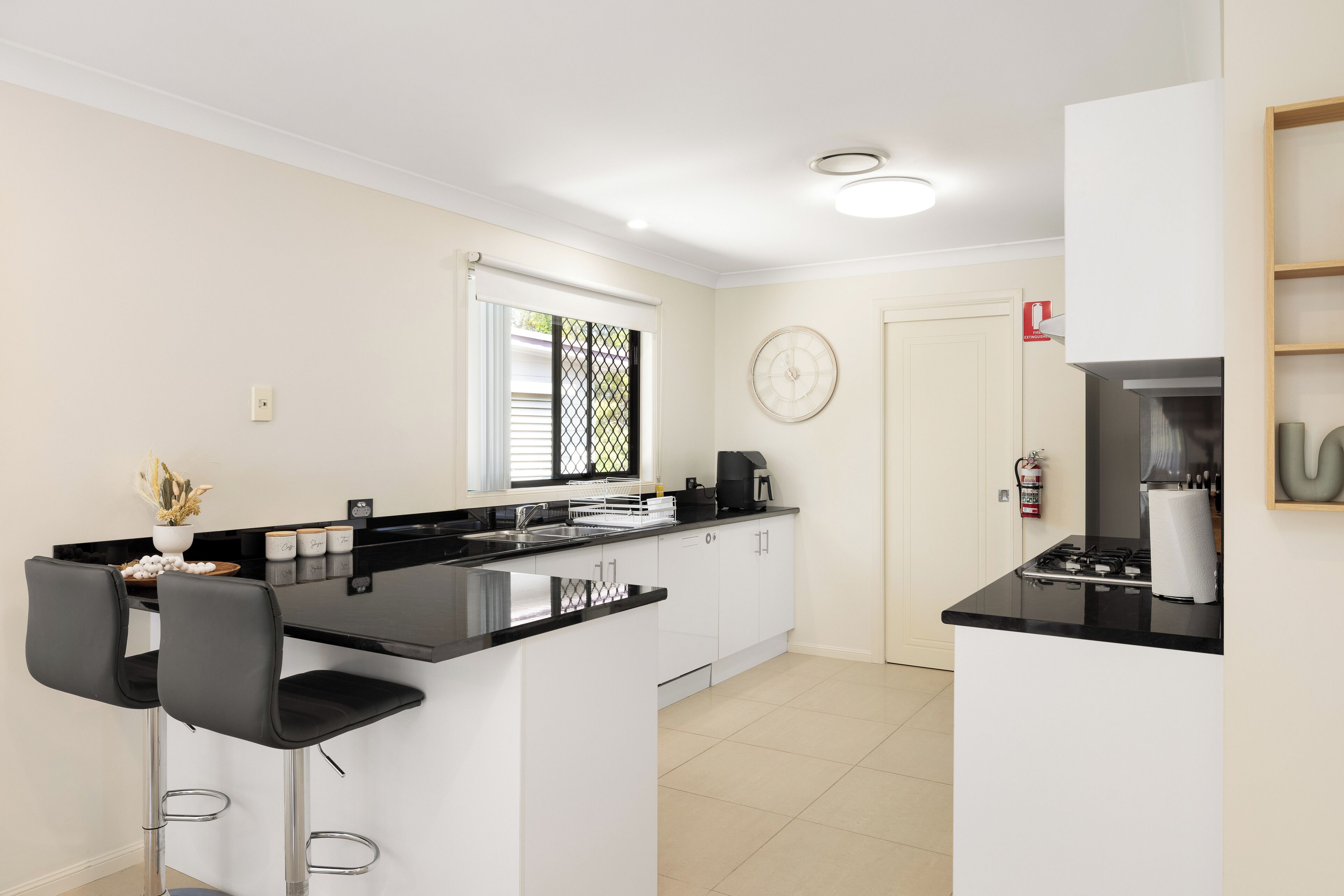 Cocina moderna con armarios blancos, encimeras negras y electrodomésticos de acero inoxidable. Hay dos taburetes de bar y una ventana con persianas.