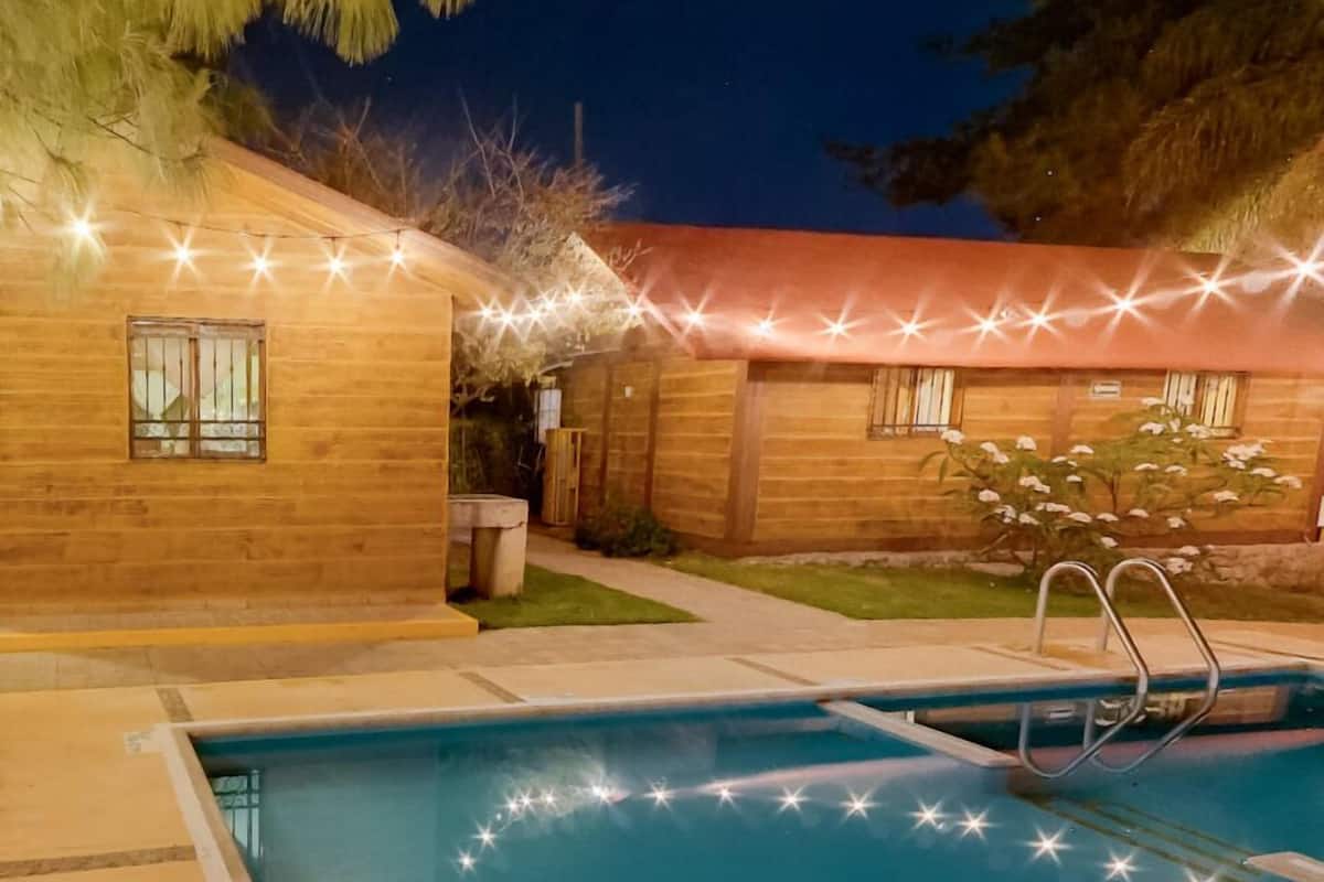 A wooden house with a swimming pool, surrounded by a well-lit garden at night.