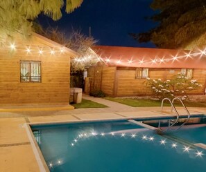 A wooden house with a swimming pool, surrounded by a well-lit garden at night. - Parque Girasoles Balneario & Cabañas (Purísima del Rincón)