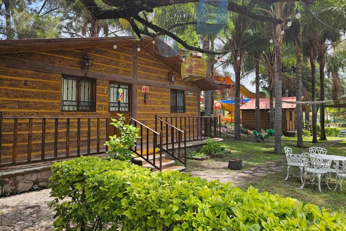 A rustic wooden cabin with a balcony, surrounded by lush greenery and a playground.