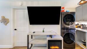 A modern living room with a flat-screen TV mounted on a wall, a built-in washer and dryer, a fireplace, and a wooden floor. - Charming 1-Bedroom Guest House Near NRG with a Level 2 Fast EV Charger (Houston)