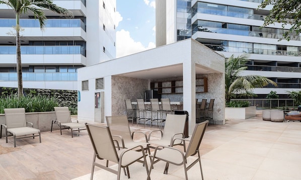 Un patio al aire libre con suelo de losas de piedra, mobiliario exterior con sillas y mesas de metal, y vistas de un edificio moderno con varios balcones.