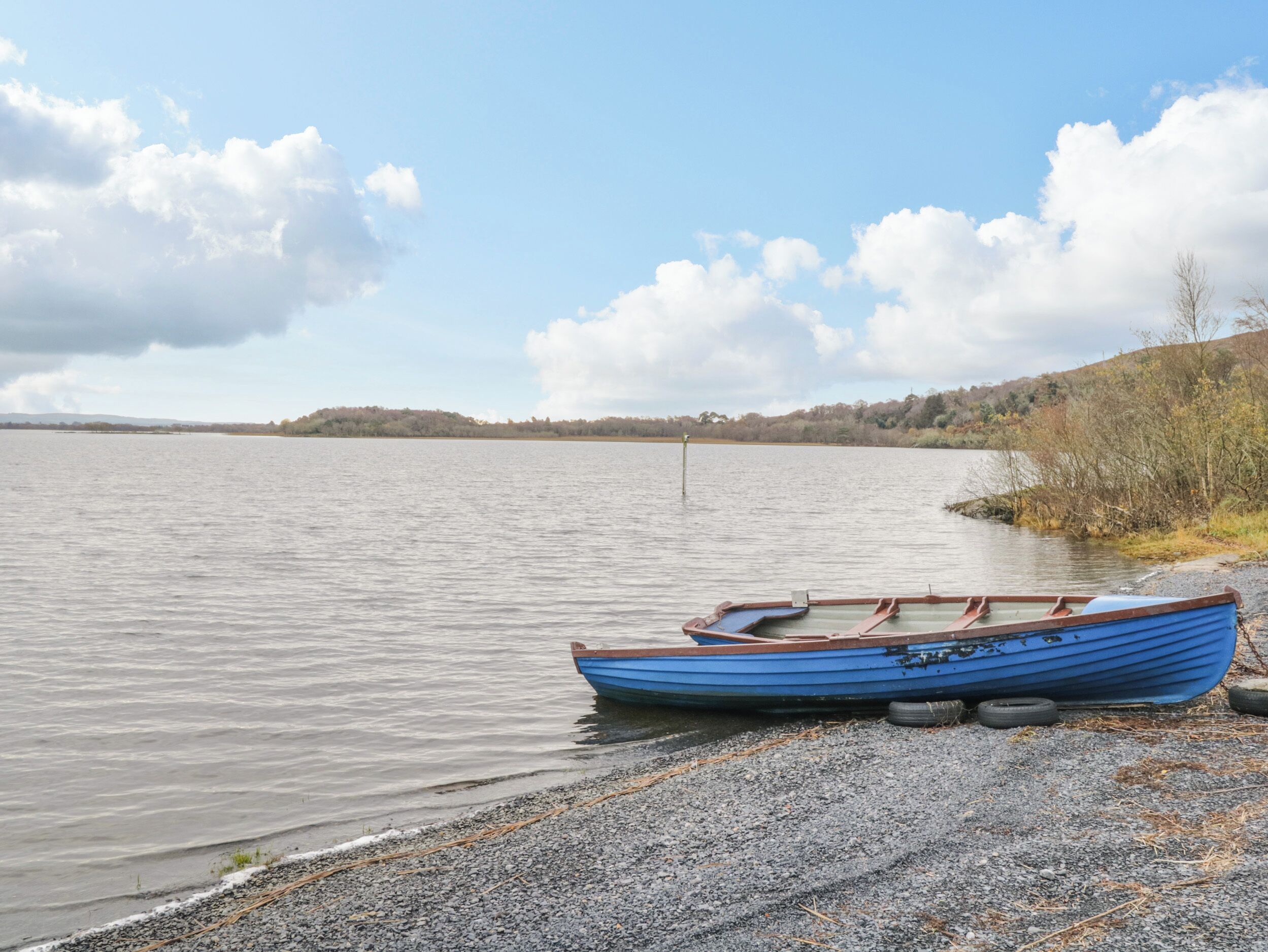 A blue rowboat on a pebbled shore by a calm lake with a distant shoreline and trees.