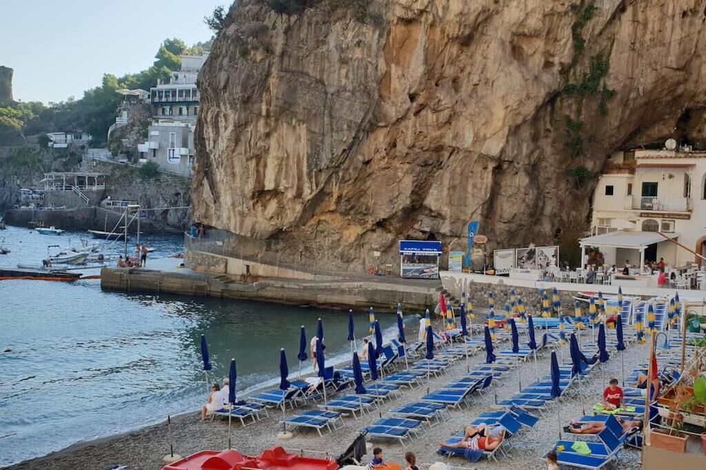 Una playa costera con hileras de tumbonas y sombrillas, un acantilado rocoso y edificios al fondo.