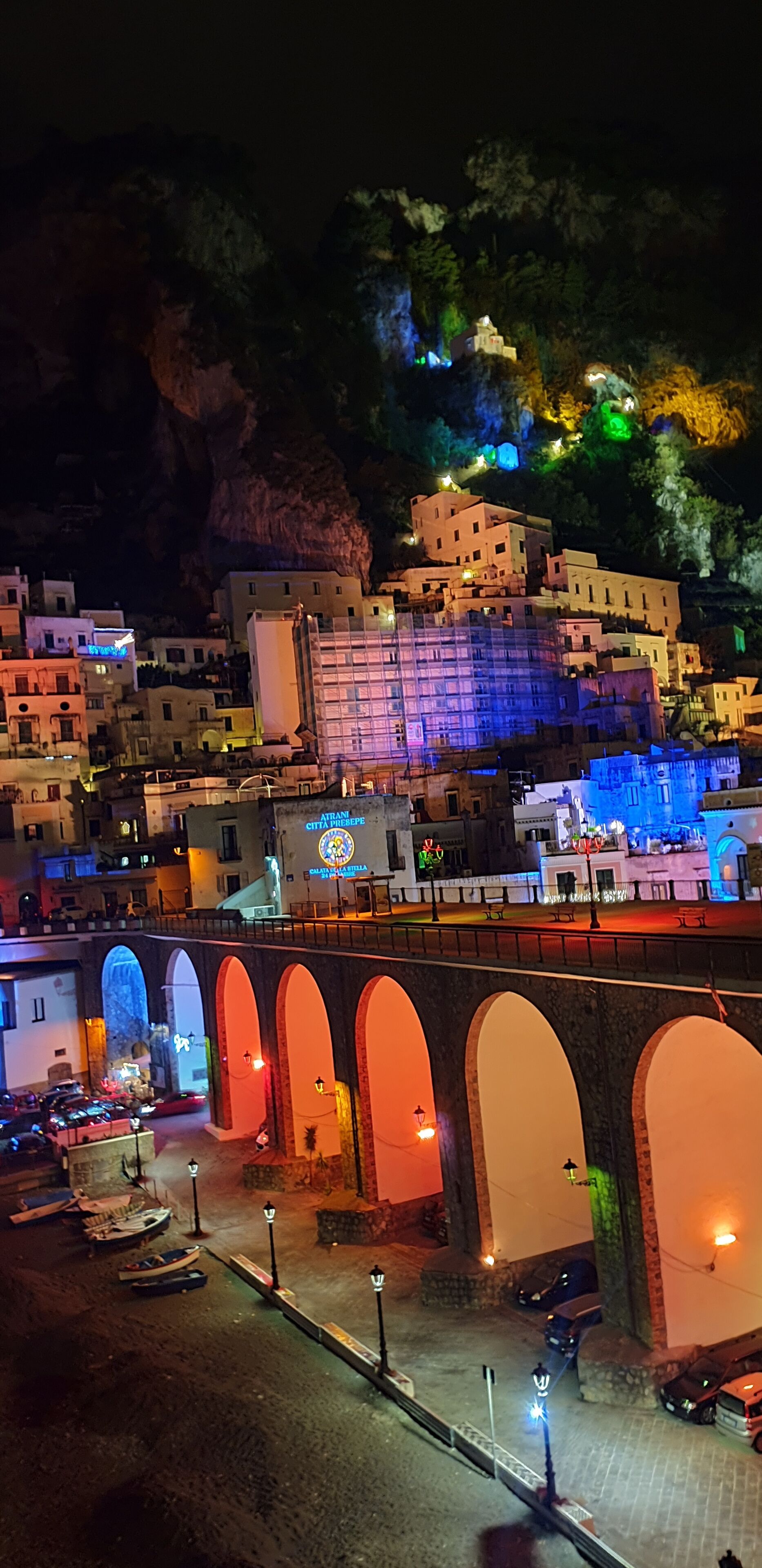 Vista nocturna de un pueblo con un puente destacado que tiene pasajes arqueados y una fila de coches estacionados.