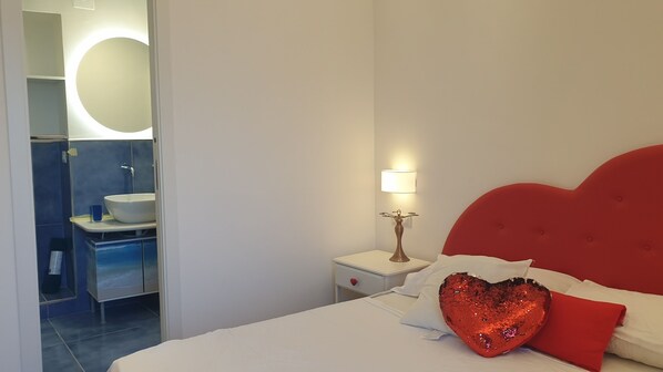 Un dormitorio con cama, mesita de noche, un edredón y un baño con lavamanos y espejo.