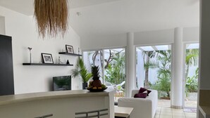 Ein moderner Wohnraum mit Küchenzeile, einem Esstisch mit Barhocker, einem weißen Sofa und einem großen Fenster mit Blick auf tropische Pflanzen.