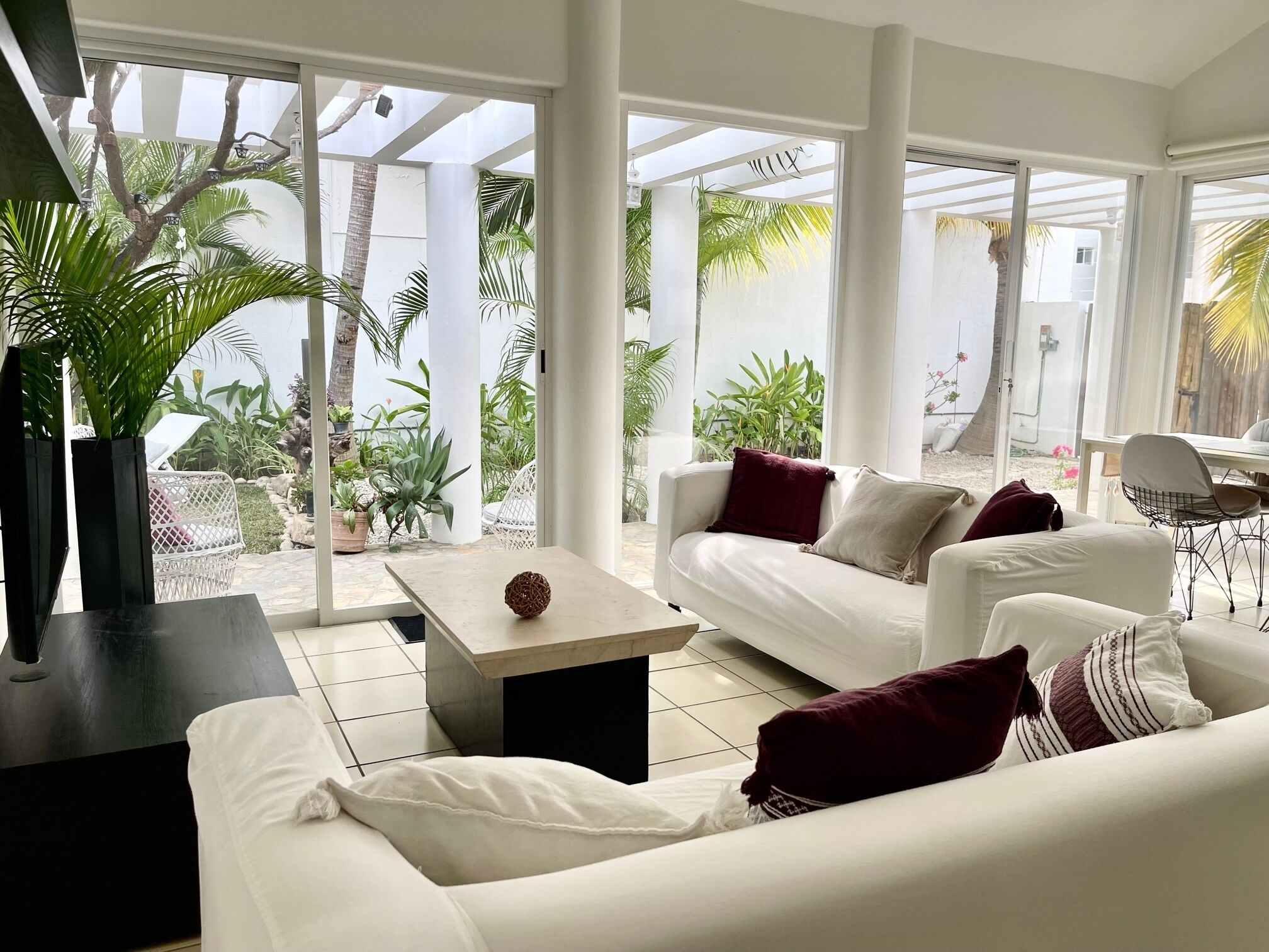 Un salón moderno con un sofá blanco, una mesa de centro y un televisor. Amplias ventanas ofrecen vistas a un jardín y palmeras.